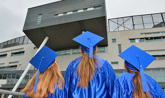 Photograph of 3 girls in blue graduation caps and robes looking up at QMU campus