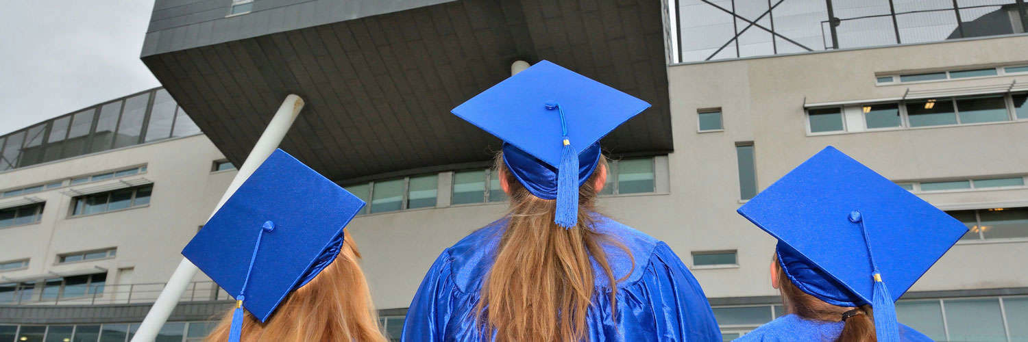 Photograph of 3 girls in blue graduation caps and robes looking up at QMU campus