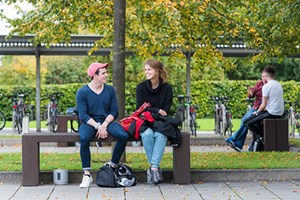 Two Queen Margaret University students sitting on a bench and chatting under some leafy green trees