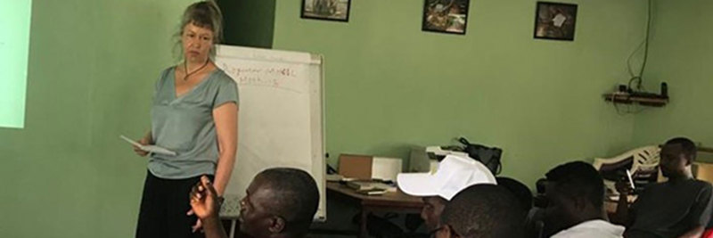 A woman standing by a projection on the wall, teaching a class of sitting men