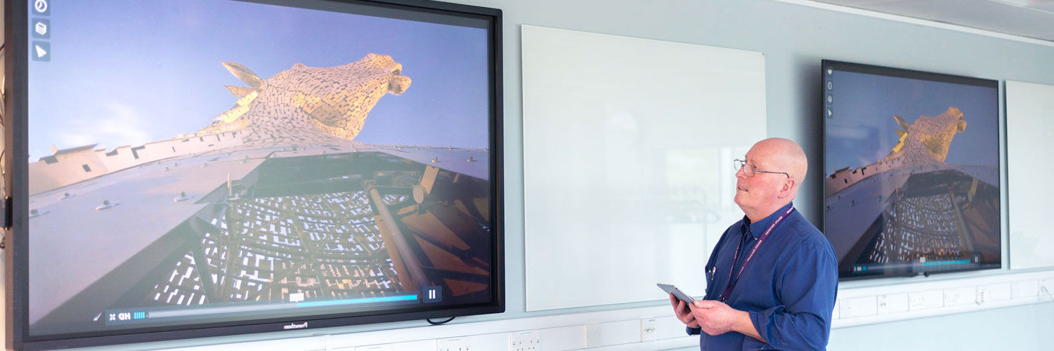 A QMU staff member standing by 2 screens showing the Scottish Kelpies sculpture