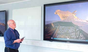 A QMU staff member standing by 2 screens showing the Scottish Kelpies sculpture