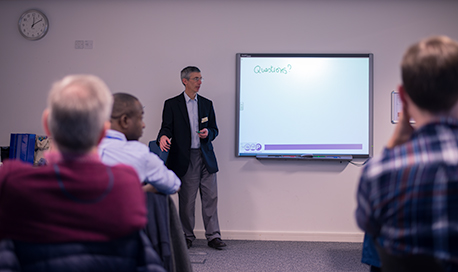 Photo taken from the back of the room during a presentation, the board says "questions?"