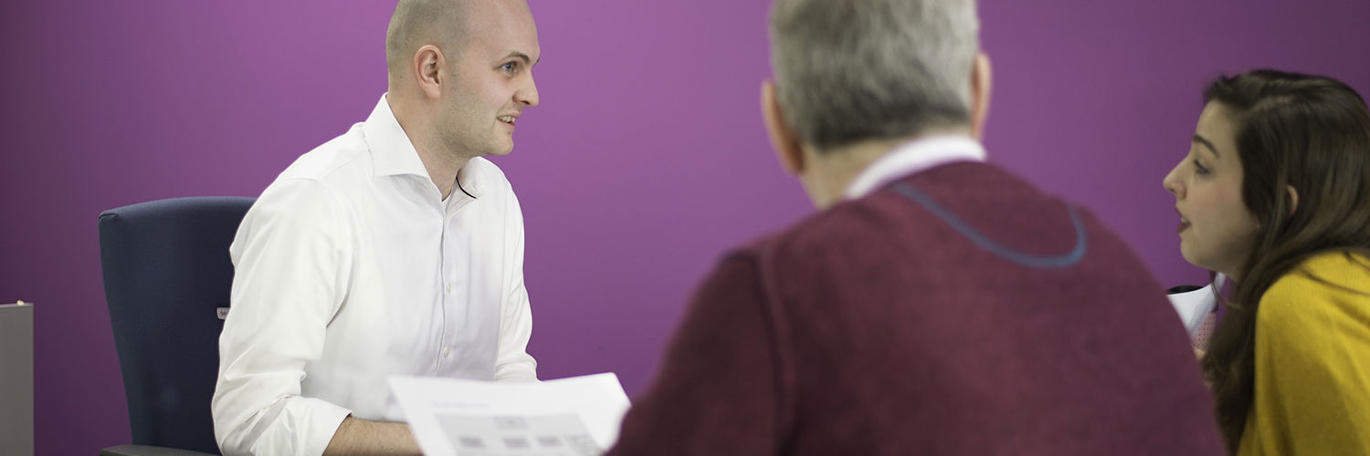 Three people at a desk talking in front of a bright pink wall