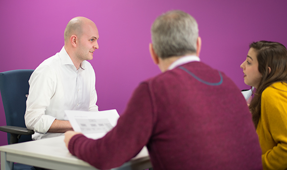 Three people at a desk talking in front of a bright pink wall
