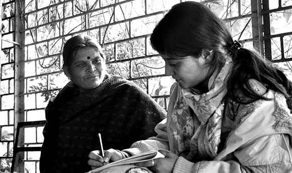 Black and white photograph of a Hindu woman talking to a younger woman taking notes