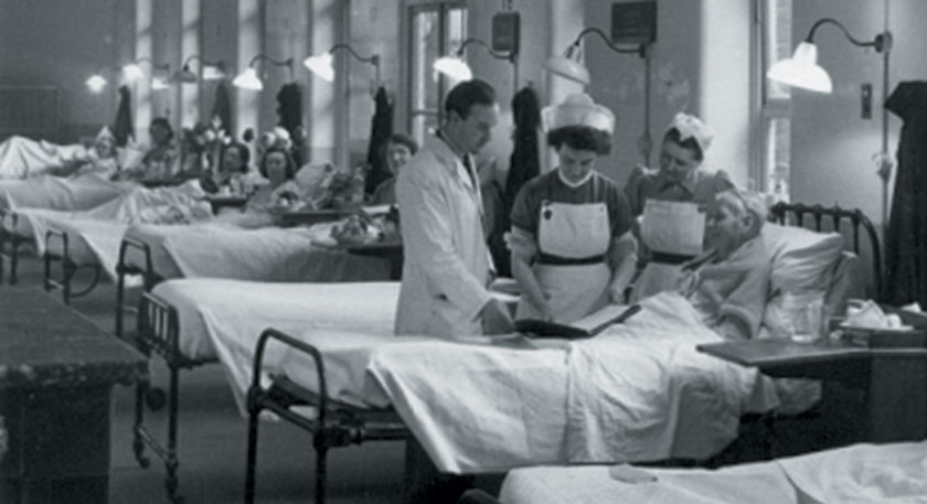 B+W photo of a hospital ward full of patients in beds with a doctor and nurses tending them
