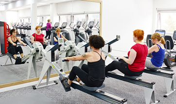 A row of women using rowing machines in front of a mirrored wall, QMU