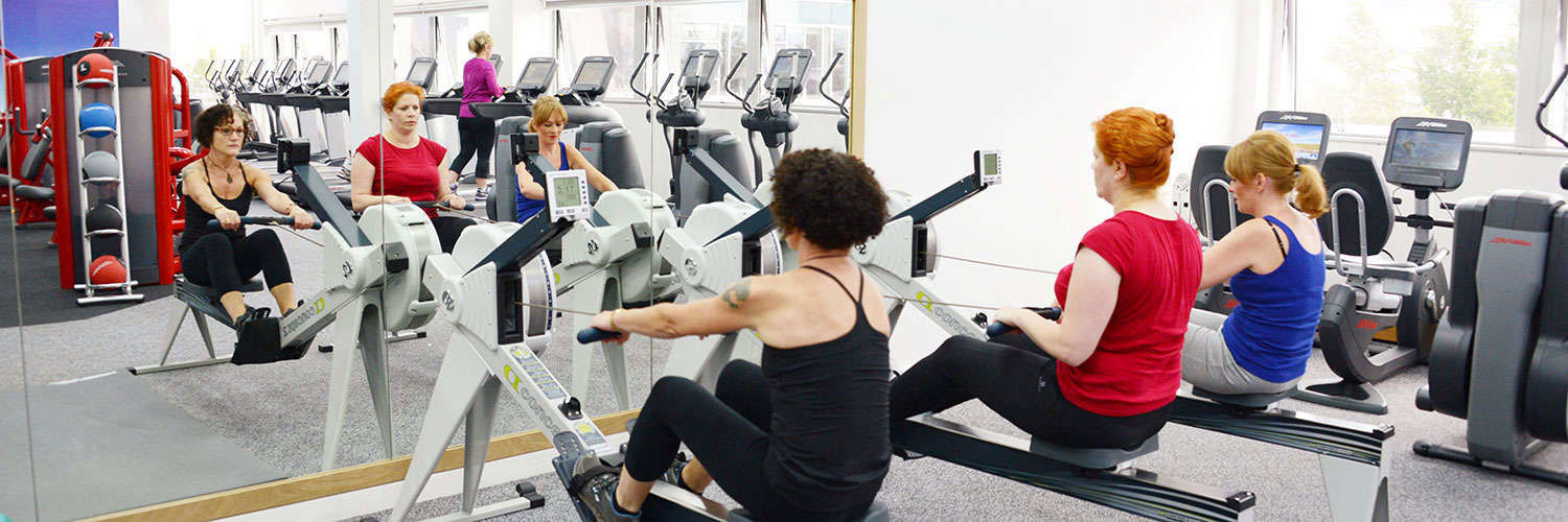 A row of women using rowing machines in front of a mirrored wall, QMU
