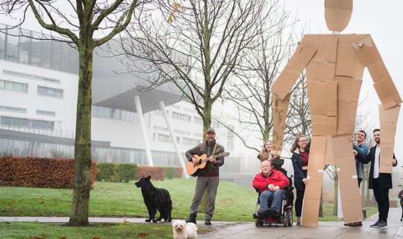 Students outside QMU holding a giant card board person up whilst a man with a dog plays guitar