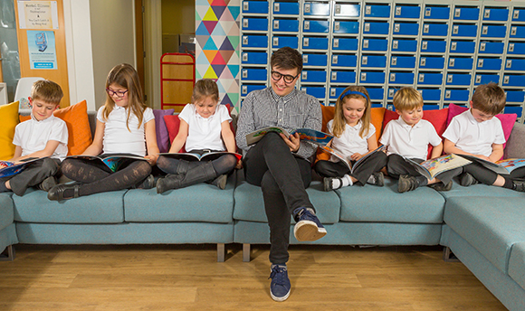 Student teacher on a sofa with 6 children reading a book together