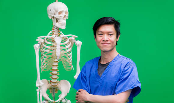Man in scrubs standing next to a model skeleton 