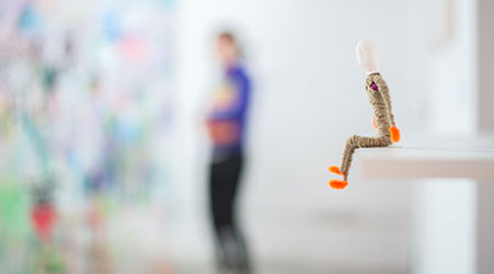 A figure of a person made of pipe cleaners and yarn sitting on the edge of a shelf