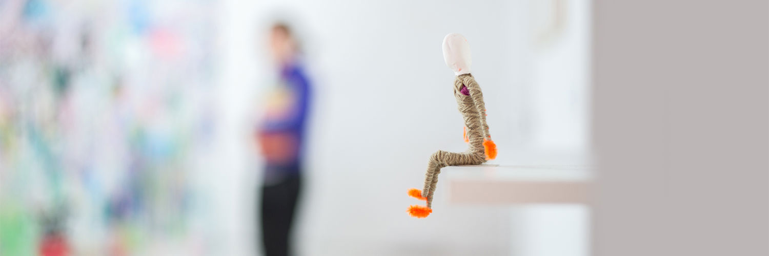 A figure of a person made of pipe cleaners and yarn sitting on the edge of a shelf
