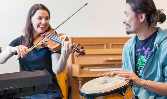 A smiling violinist and a drummer playing a bongo