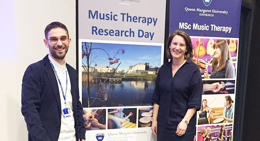 Two smiling lecturers standing by a Music Therapy Research Day sign