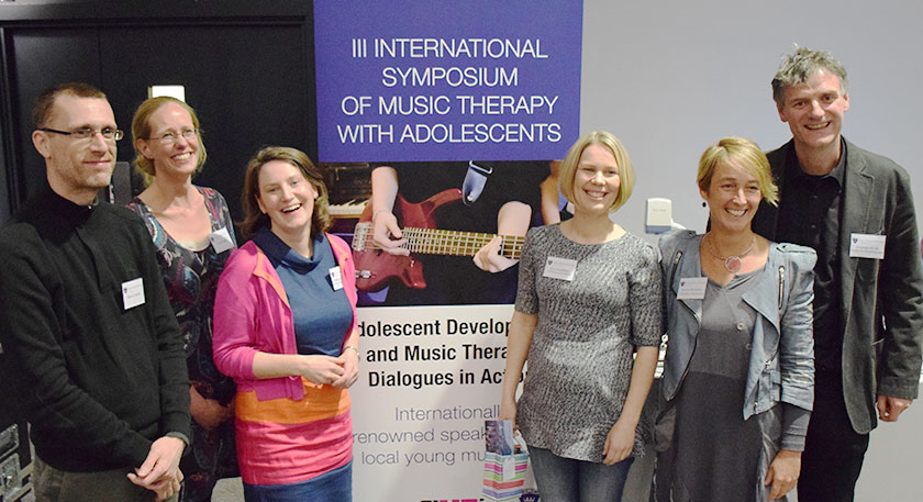 A group of smiling people beside a sign for the International Symposium of Music Therapy With Adolescents