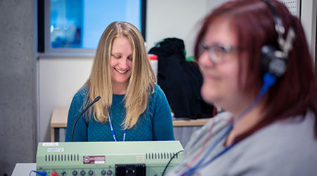 A woman with a headset on and a women behind her sat at a desk