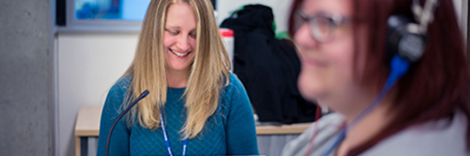 A woman with a headset on and a women behind her sat at a desk