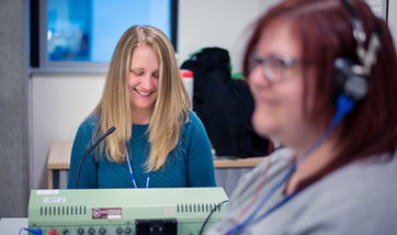 A woman with a headset on and a women behind her sat at a desk