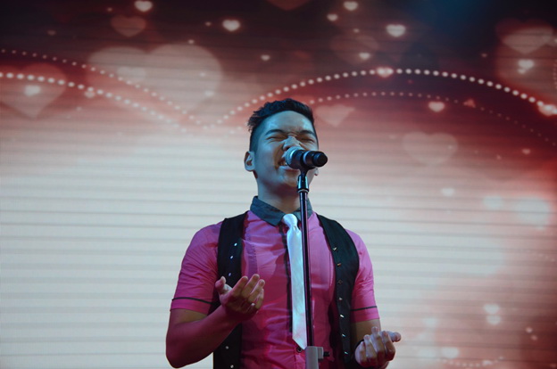 A performer on stage singing onto a mic with a backdrop covered in hearts