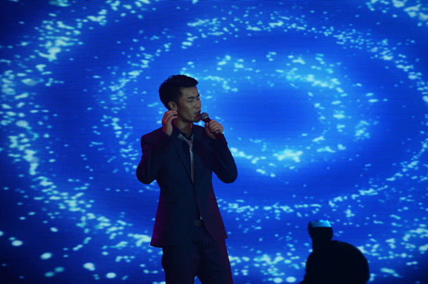 A performer singing on stage with a blue swirling back drop behind them