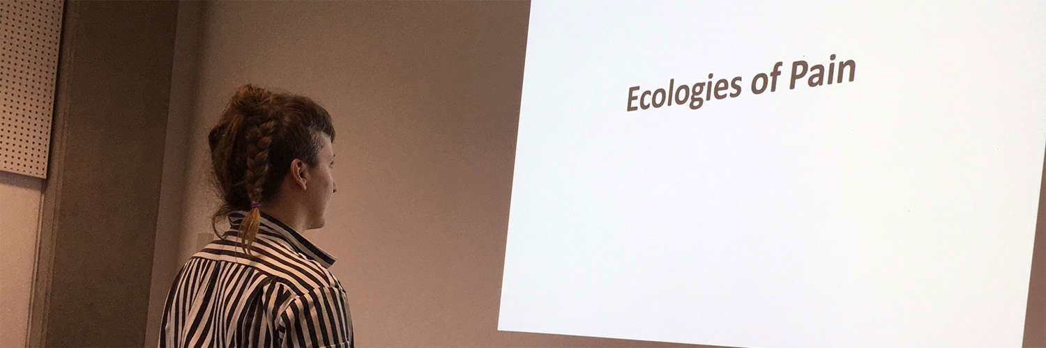 A student stands in front of a projection of the words "Ecologies of Pain"