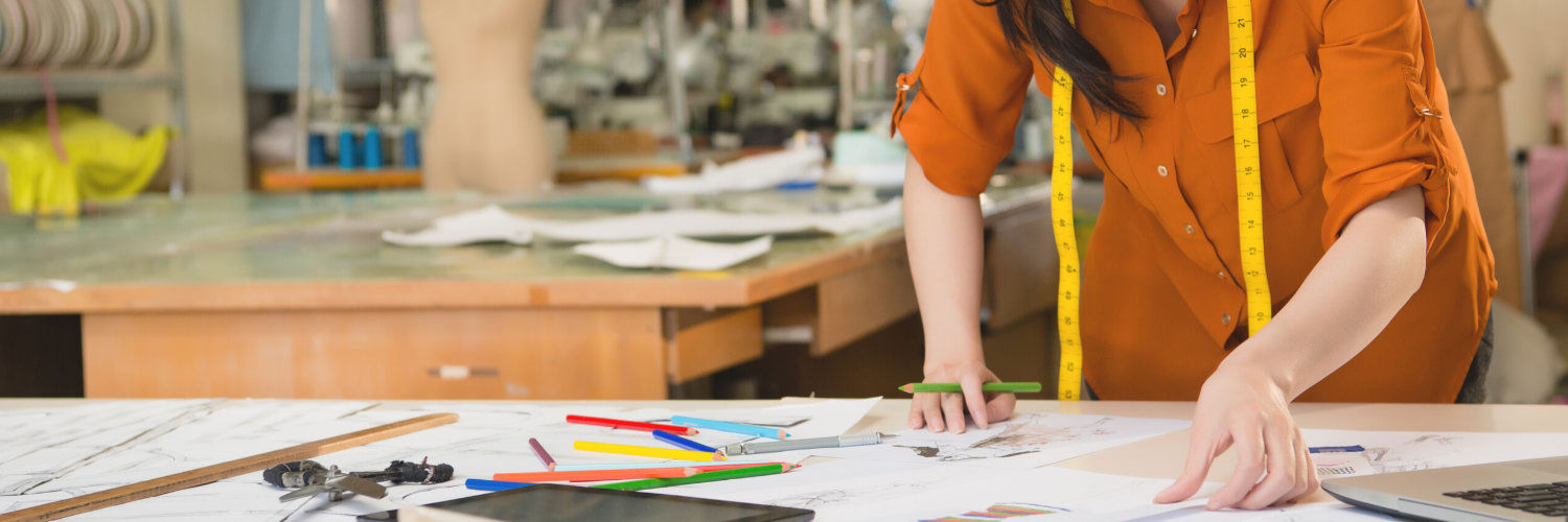 A table covered in paper, pencils and scissors stood over by a person with a tape measure round their neck