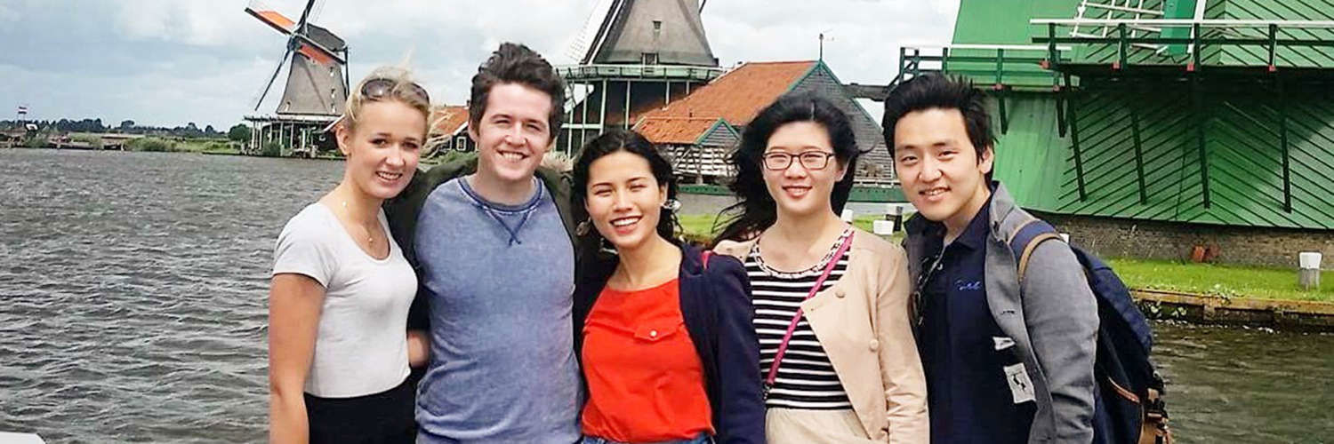 A row of smiling students posing for a photo by some windmills in Holland