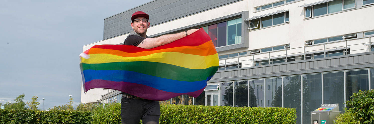 A QMU student holding up rainbow LGBTQIA+ flag in front of campus building