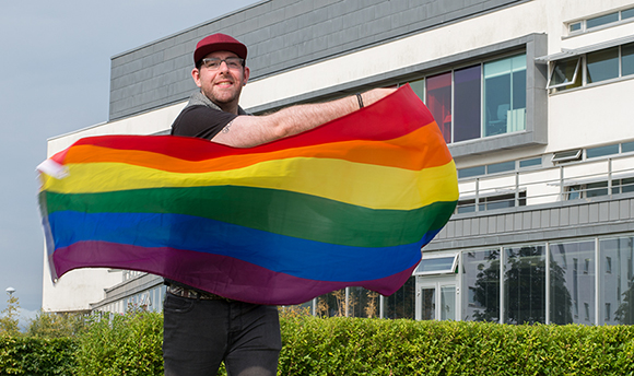 A young man smiling outside QMU and holding up the rainbow LGBTQIA+ flag