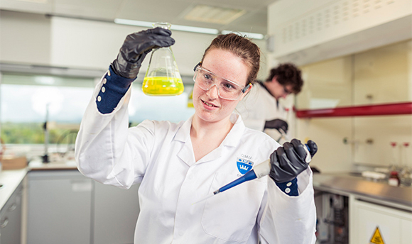 A QMU student wearing PPE and holding up a beaker of yellow liquid