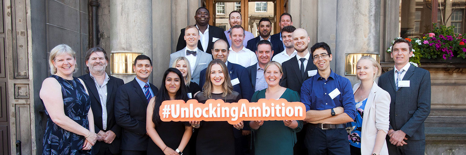 A large group of well dressed people standing on steps and holding a sign that reads '#UnlockingAmbition'