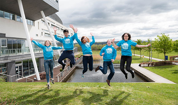 A group of QMU students in matching blue sweaters on campus jumping together for the photo