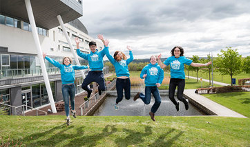A group of QMU students in matching blue sweaters on campus jumping together for the photo