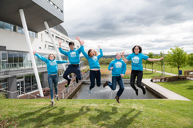 A group of students in matching blue sweaters jumping for a photo outside QMU