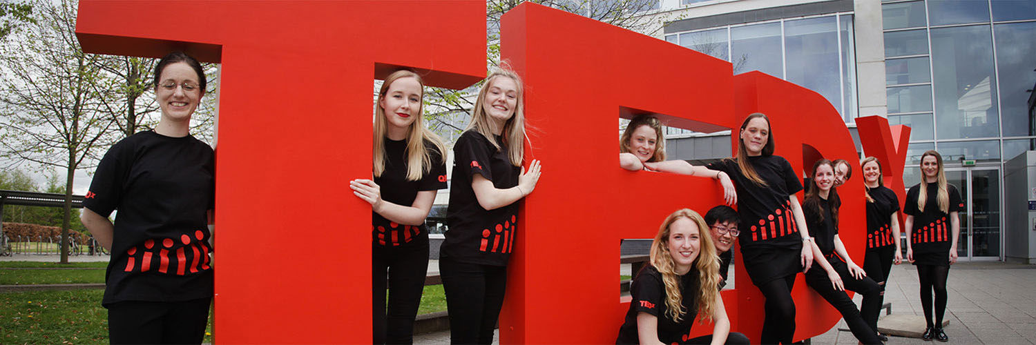 A group of young women in matching shirts posing for a photo by the large TEDx sign