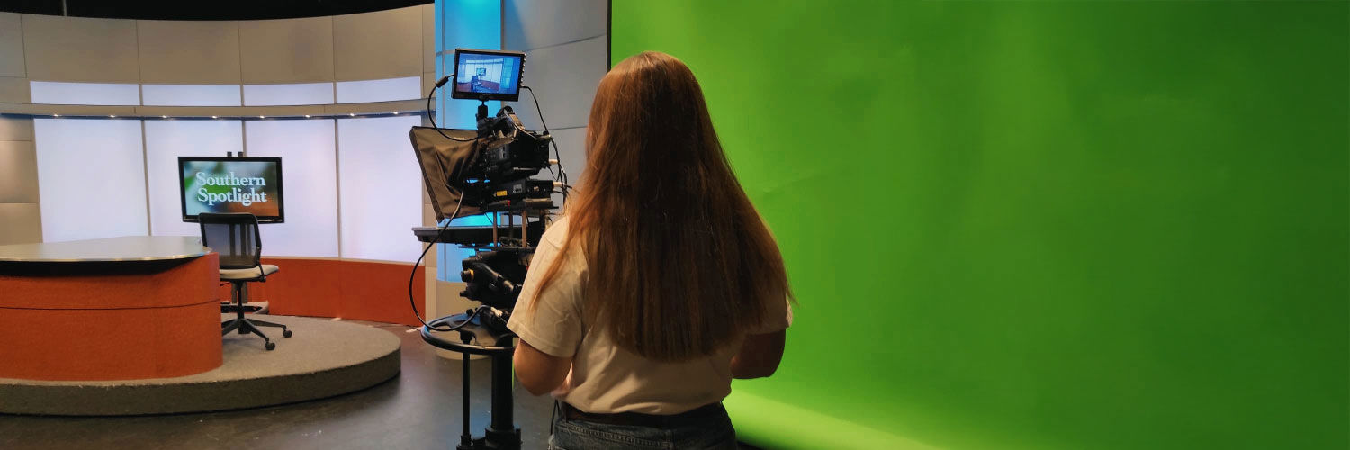 A young woman operates a film camera inside what appears to be a news studio.