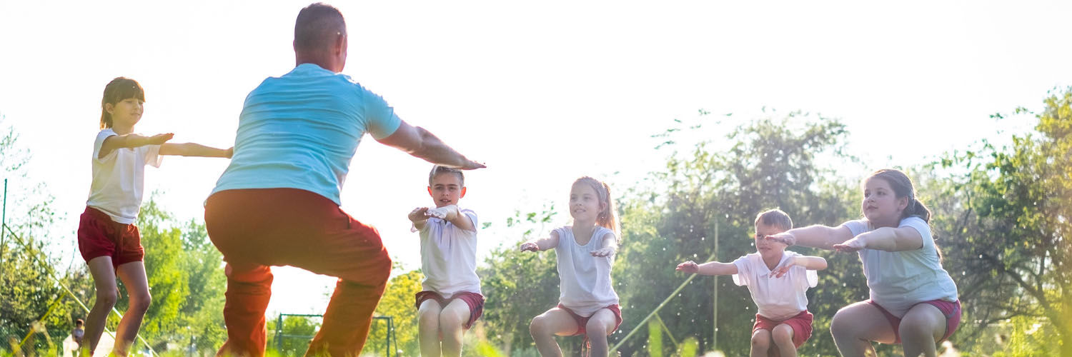 A teacher and a group of five children outside in a park performing squats.