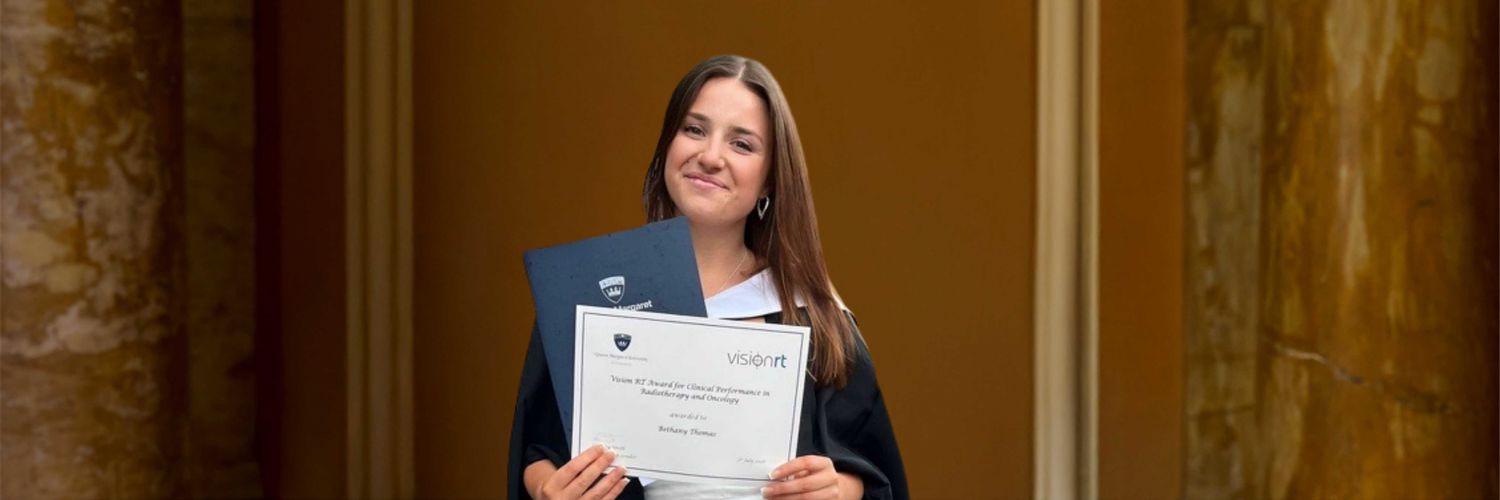 head and shoulders shot of female student with long brown hair in graduation robes and holding up certificate