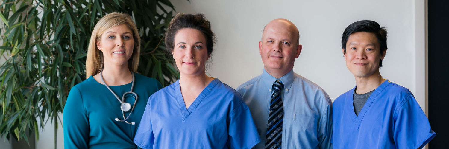 Four medical practitioners wearing blue scrubs stand smiling at the camera against a plain wall, with a large plant in the background.