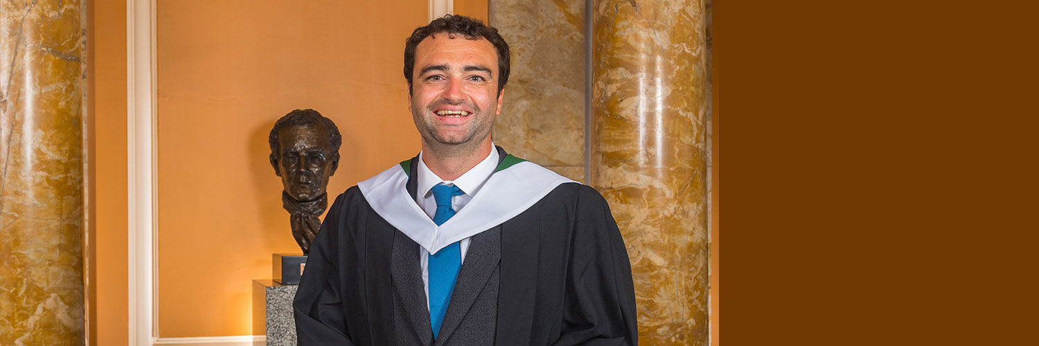 Portrait shot of Stewart Grant in his graduation robes holding his graduation scroll