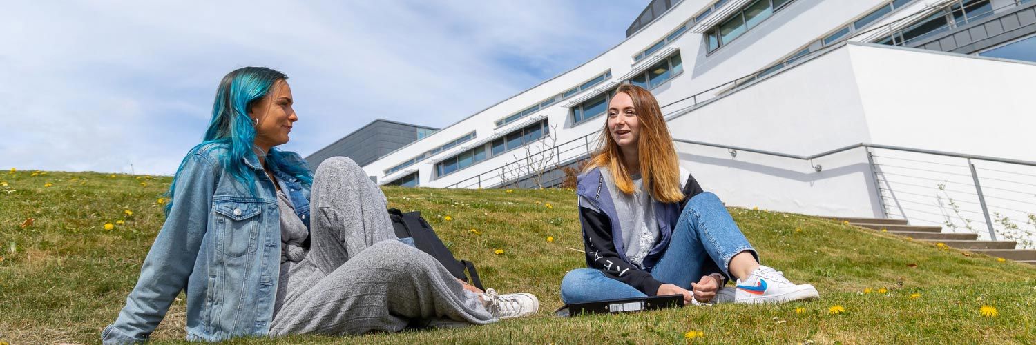 Two young women sit on the grass outside the main QMU building