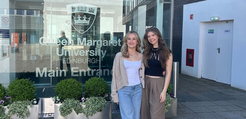 Two students standing in front of the main QMU campus entrance.