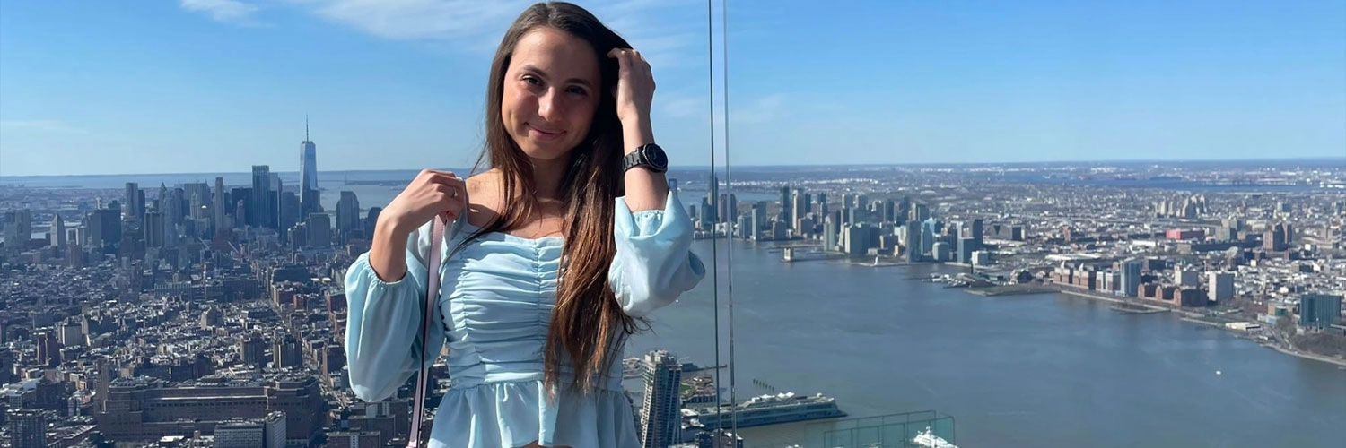 A young woman poses for a photo from the top of a skyscraper, the view of the cityscape and intersecting river are seen behind her.