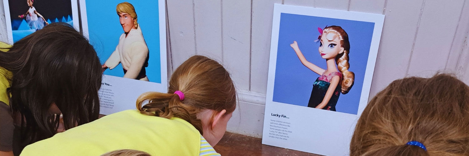 A group of children drawing on pieces of paper, some images of popular dolls and toys with visible limb differences are on display.