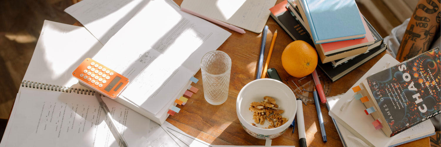A table scattered with different books, notepads, stationery and snacks.