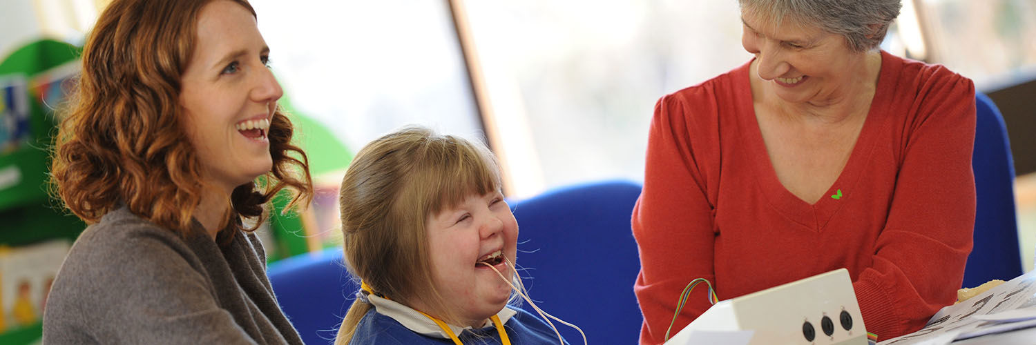 A young girl with Downs Syndrome using the speech and language therapy equipment with two adults
