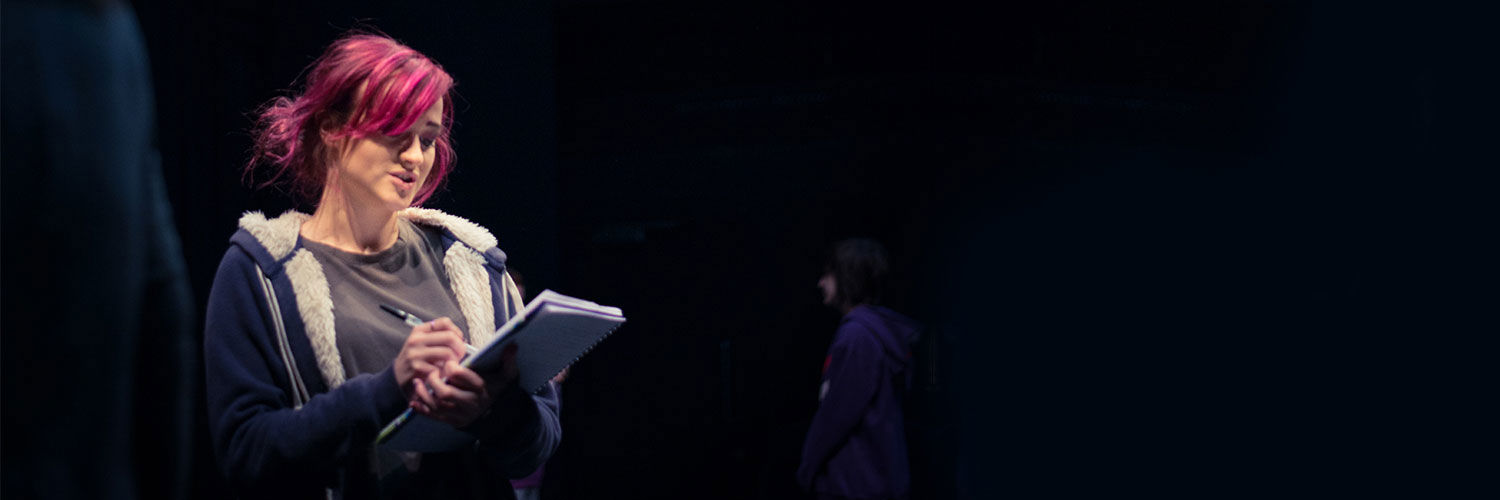 A student with bright pink hair stands on stage reading out instructions from a clipboard.