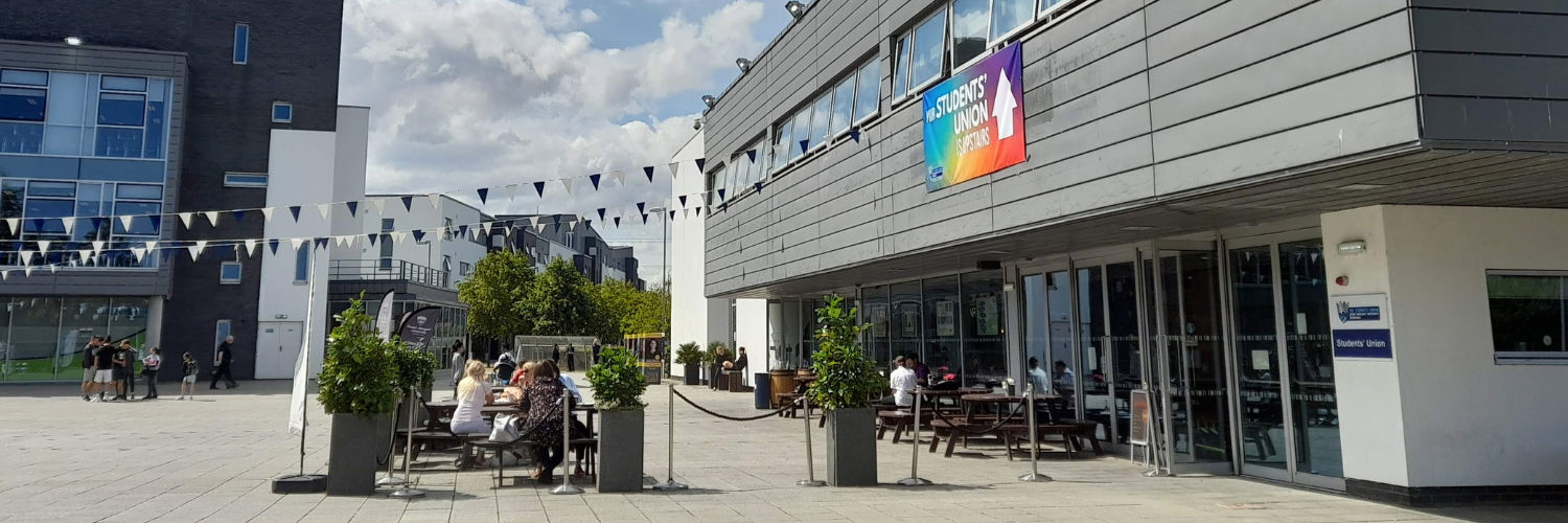 Groups of people sitting at picnic tables outside a cafe on a university campus building, on a sunny day.
